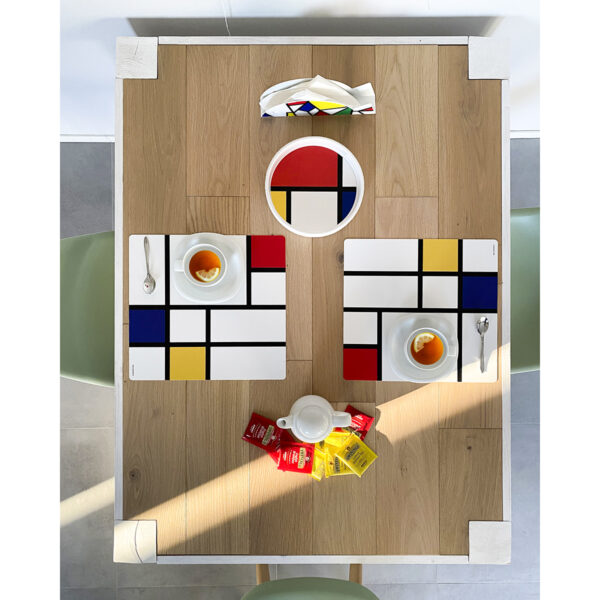una tavola in legno è apparecchiata con due tovagliette americane quadrate, una ciotola portapane e un portatovaglioli tutti nello stile artistico Mondrian