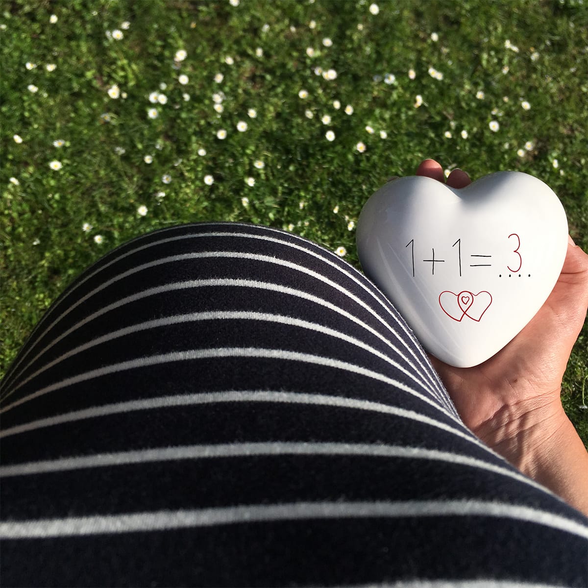 un cuore con la scritta 1+1=3 è tenuto in mano vicino ad una pancia di una futura mamma in attesa