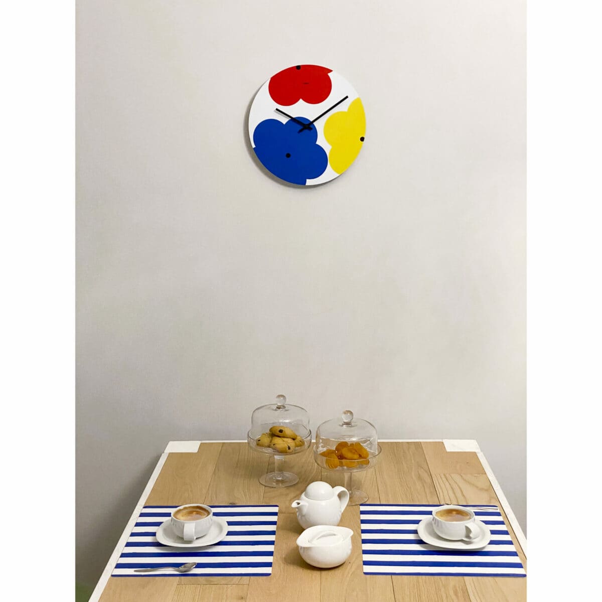 sopra un tavolo in legno apparecchiato per la colazione è appeso un orologio a muro di ispirazione pop, rotondo, raffigurante 3 maxi fiori stilizzati nei colori rosso, blu e giallo