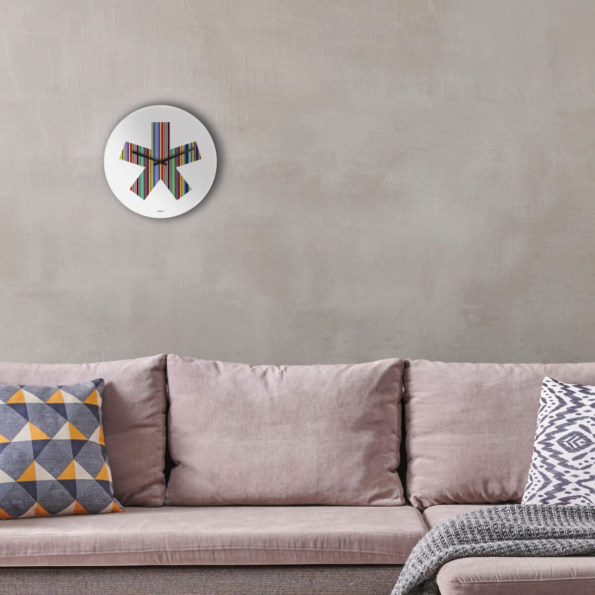 sopra un divano color tortora è stato appeso un orologio a muro rotondo, base bianca, con la stampa di un grande asterisco a 5 braccia multirighe e multicolore