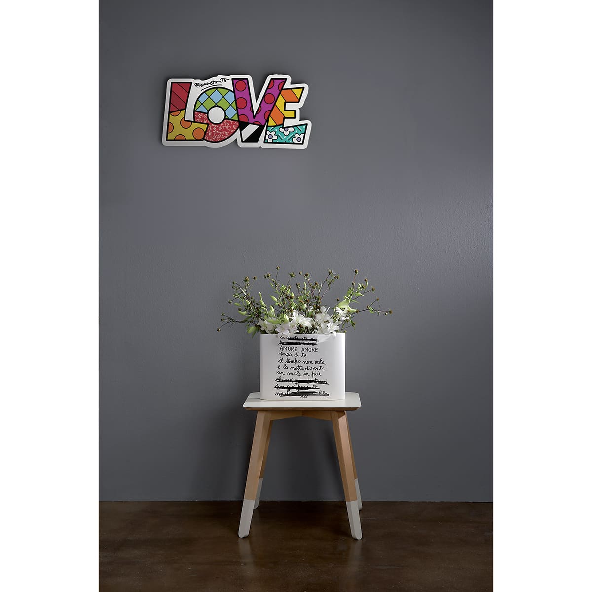 la scritta "love" in stile street art è appesa come elemento decorativo su una parete grigia, sopra ad un tavolino con un vaso di fiori