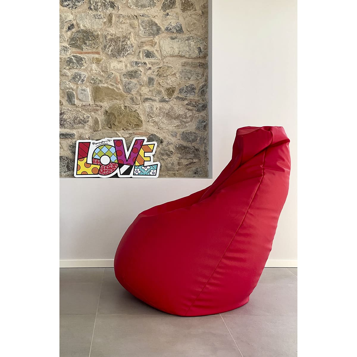 un pannello decorativo con la scritta pop "Love" è appoggiato sulla rientranza di un muro di pietra vicino ad una poltrona sacco rossa