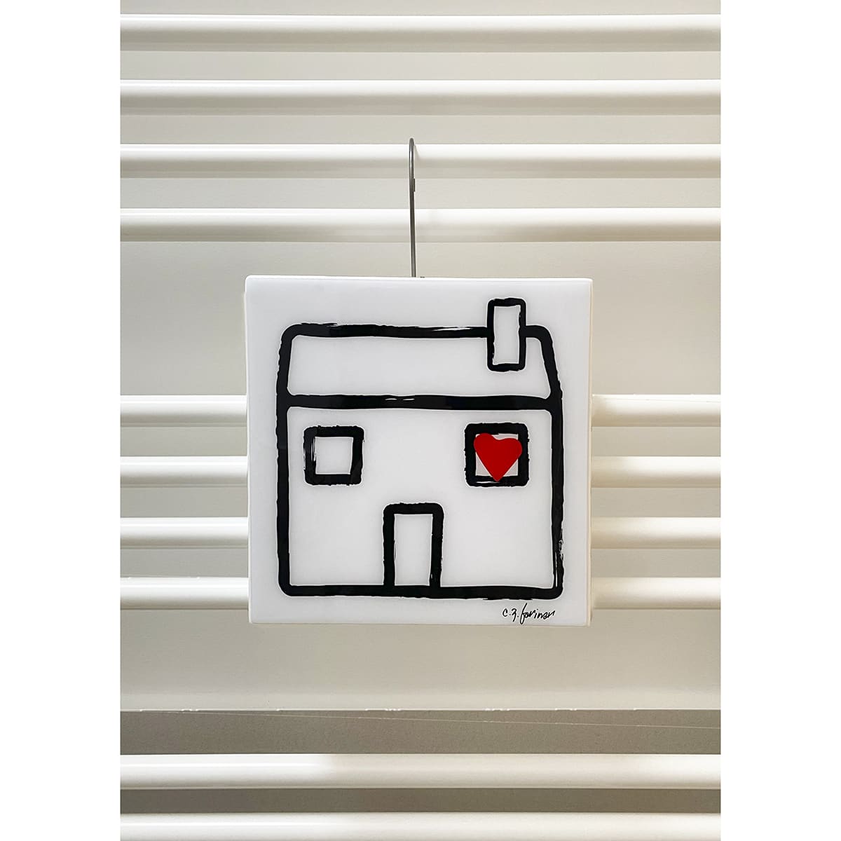un umidificatore quadrato con il disegno in nero di una casa stilizzata dalla cui finestra esce un cuore rosso è appeso ad uno dei tubi di un radiatore bianco