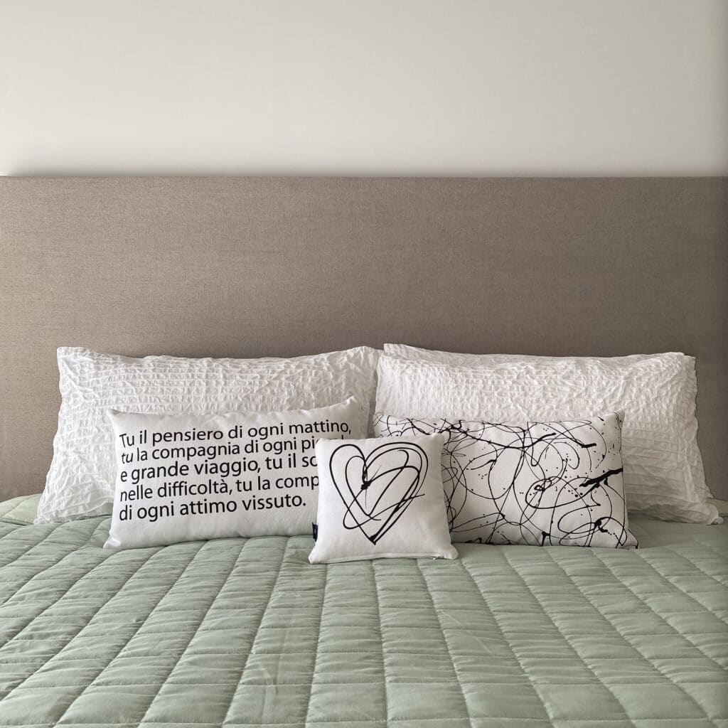 testata letto grigia completa di cuscini, alcuni dei quali decorati graficamente e riportanti i versi di una poesia.