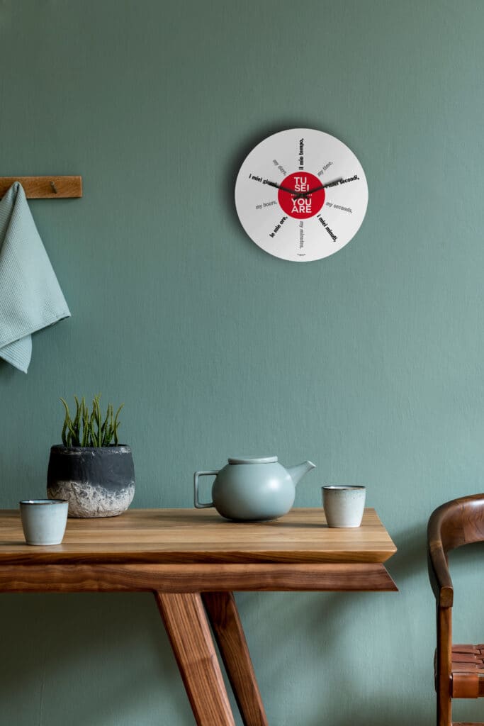 un orologio con scritte inerenti il tempo è appeso su una parete di color salvia, sopra un tavolo di legno