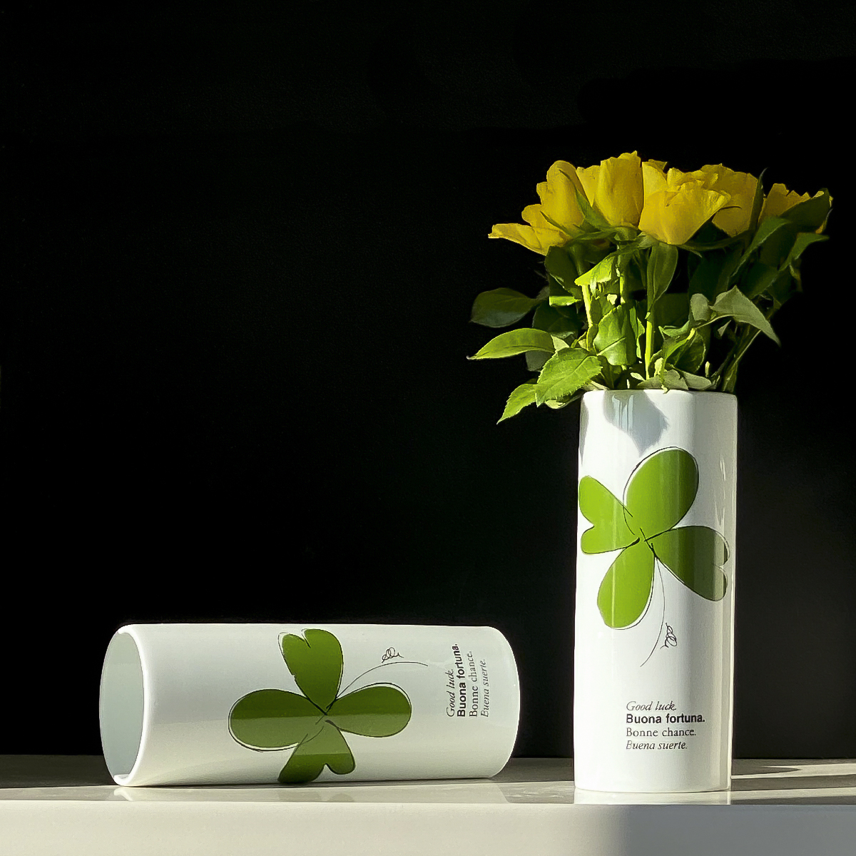 il vaso cilindrico bianco con artwork di un quadrifoglio e scritta portafortuna è fotografato con un pò di roselline gialle al suo interno.